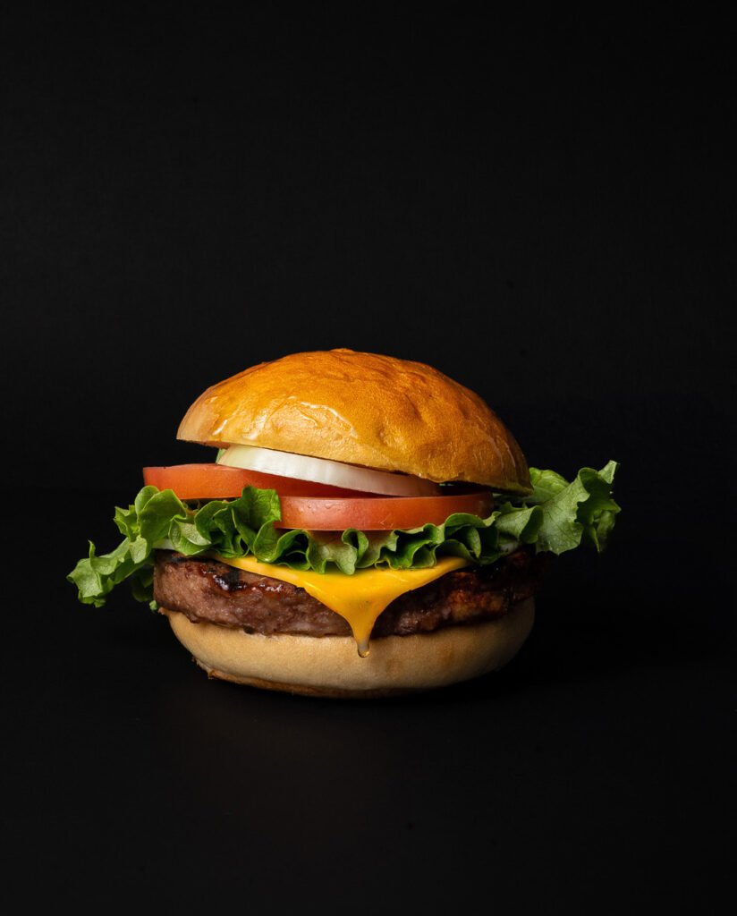 A cheeseburger with cheddar cheese, lettuce, tomato, onion, and a beef patty in a golden bun, captured by a toronto-food-photographer against a plain black background.