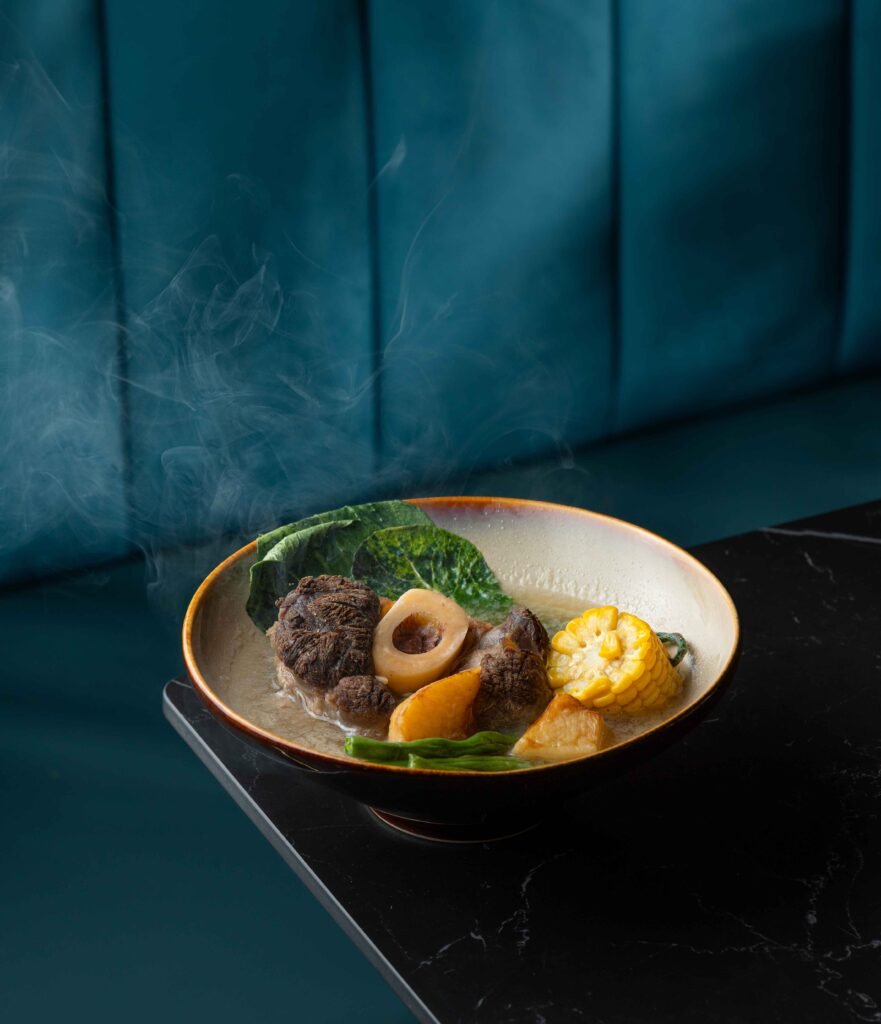 A steaming bowl of soup with bone marrow, corn, leafy greens, mushrooms, and veggies sits on a dark marble table—perfect content for a social-media-agency to showcase against a stunning teal backdrop.