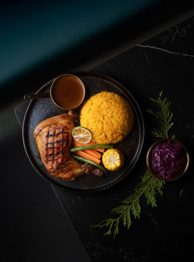 A black plate with grilled chicken, yellow rice, grilled vegetables, a slice of lime, corn, brown sauce, and purple cabbage garnished with green leaves—perfect for those seeking a GTA premium dining experience on a dark surface.