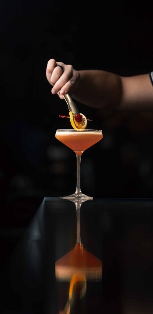 A professional drink photographer captures a dramatic moment as a hand garnishes an orange cocktail with a dried orange slice and cherry, its reflection shimmering on the glossy surface beneath the cinematic, dark backdrop.
