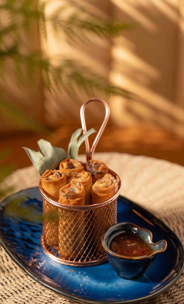 A small basket filled with crispy spring rolls sits on a blue plate, accompanied by a small bowl of dipping sauce—a perfect nod to Philippines flavors. Sunlight casts shadows across the scene, creating a warm and inviting atmosphere.