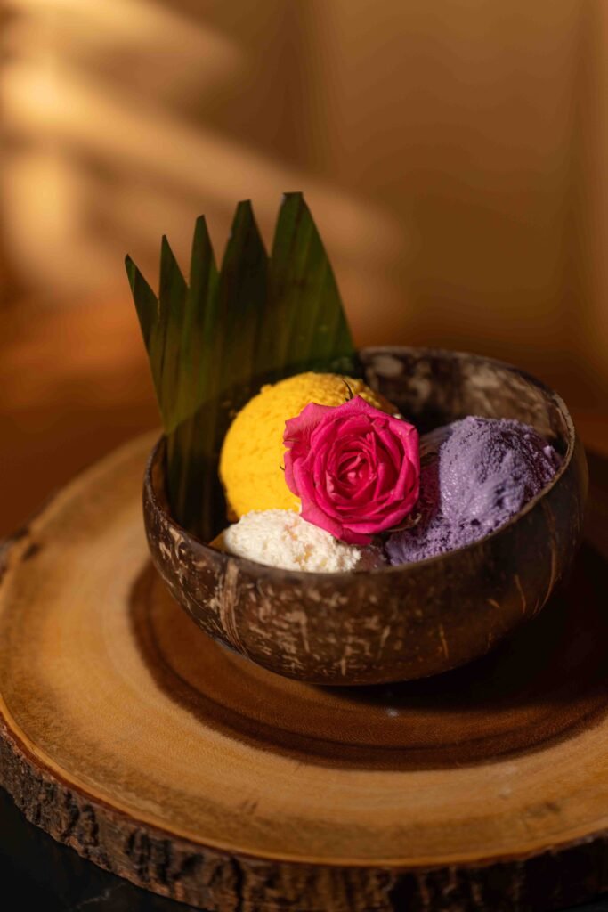 A wooden bowl with three scoops of ice cream in yellow, white, and purple, topped with a pink rose and green leaf, placed on a wooden slab with warm lighting by a talented Toronto-food-photographer.