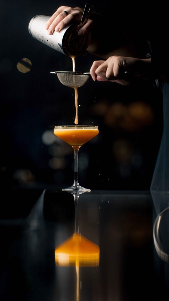 A hand pours a golden cocktail from a shaker through a strainer into a coupe glass. The drink forms a smooth stream, casting a clear reflection on the glossy black surface—a striking moment in drink photography.
