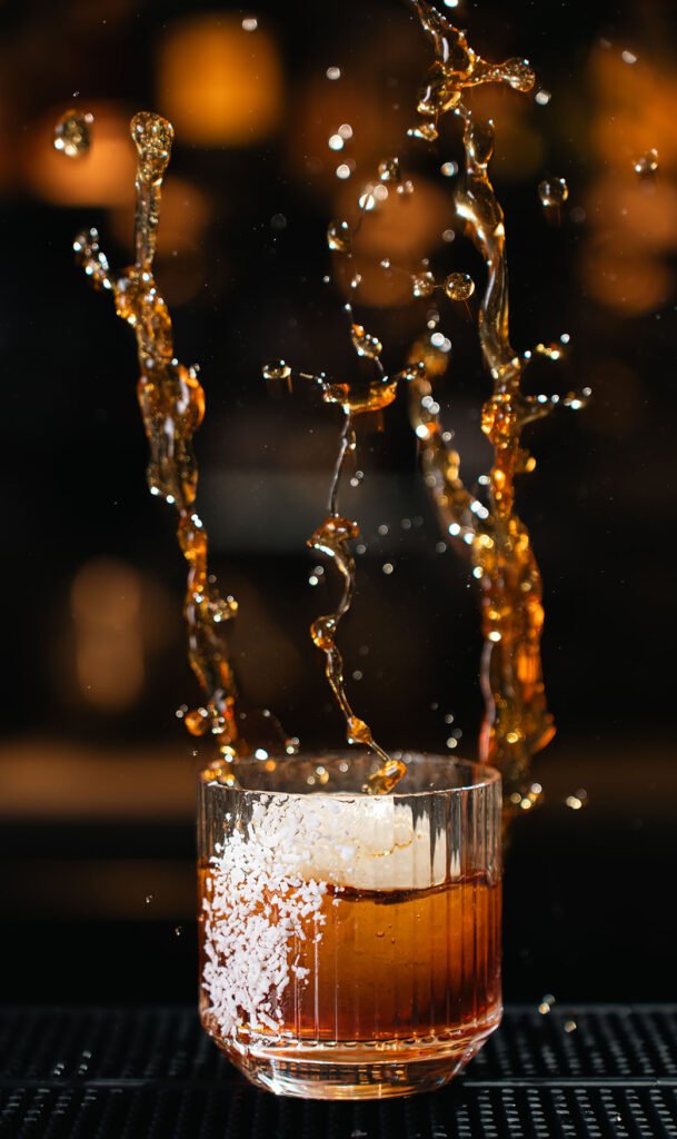 Close-up of a splashy, amber-colored cocktail with ice, illuminated by warm, soft lighting, highlighting its refreshing taste