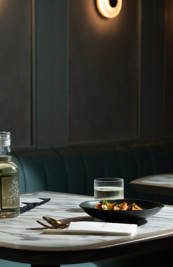 Captured by a professional-food-photographer, a marble table is set with a bottle of water, glass, cutlery, napkin, and a plate of food. In the background, a green upholstered bench and lit round wall sconce complete the scene.