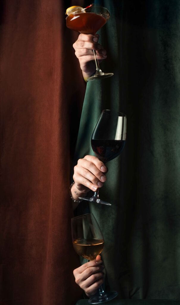 Three hands emerge from between brown and green draped curtains, each holding a different holiday drink: the top a cocktail with an orange garnish, the middle a glass of red wine, and the bottom a glass of amber liquor.