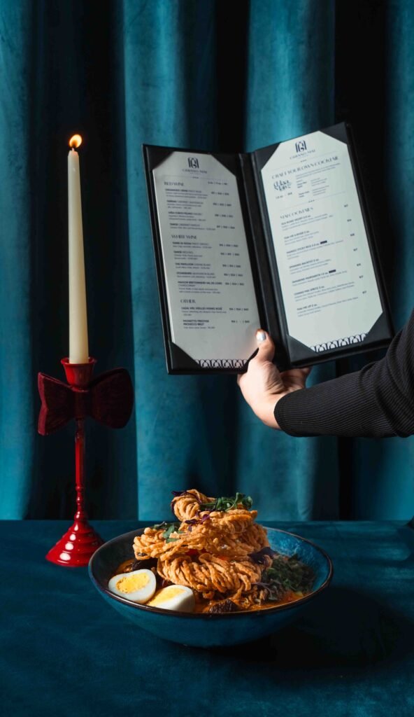 A person holds open a restaurant menu next to a lit candle in a red holder with a bow, above a table set with noodles, eggs, and garnishes. The cozy Ontario ambiance is set against a blue curtain backdrop.