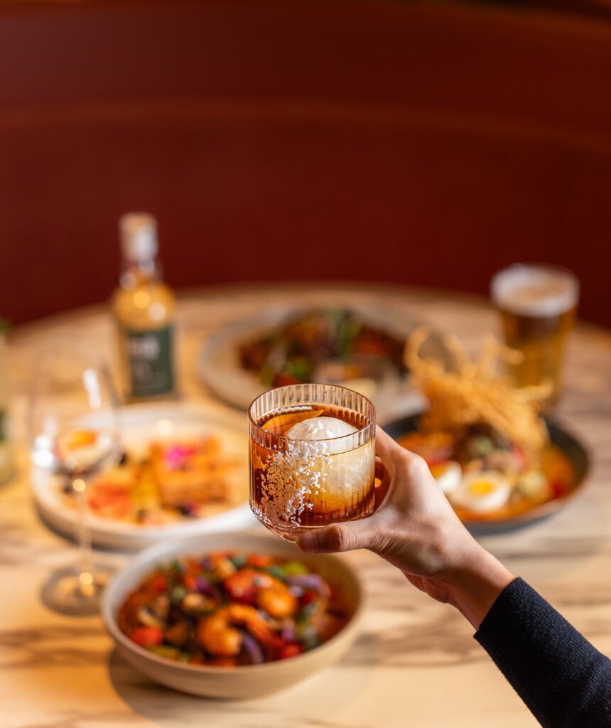 A hand holds a glass of dark cocktail with ice, in focus, while plates of food and drinks from a family dinner sit on a blurred restaurant table in the background.