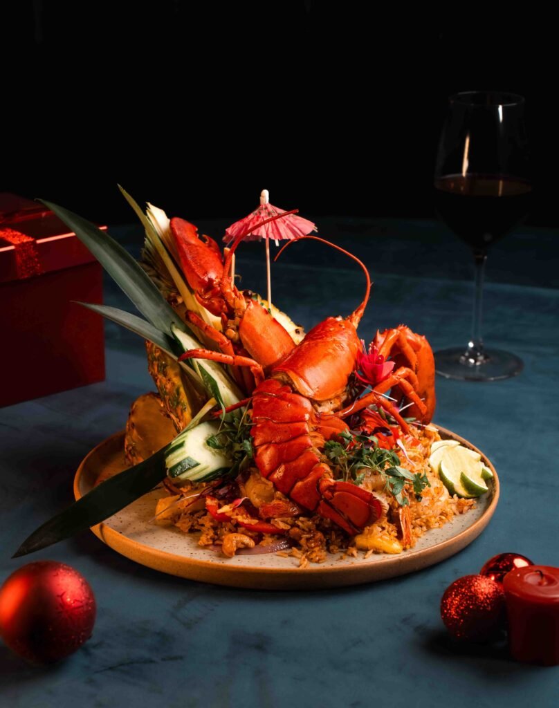 A plated lobster dish served in a halved pineapple, garnished with herbs, vegetables, and a small paper umbrella. Showcasing professional restaurant flair, it’s paired with red wine and festive ornaments on a blue table.