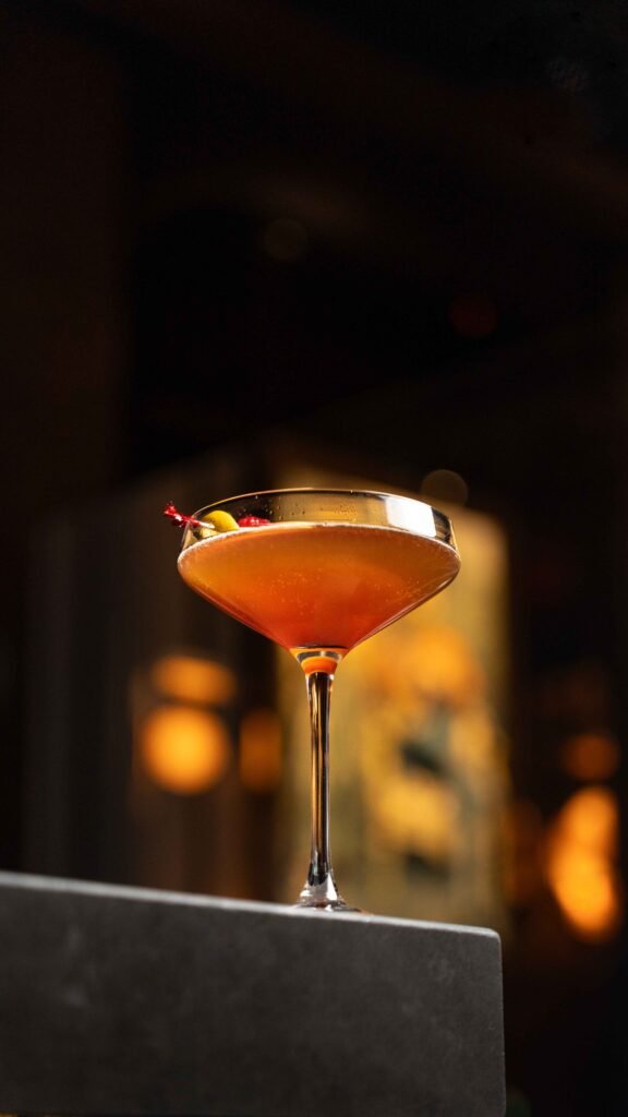 A cocktail in a coupe glass garnished with a cherry and a twist of citrus peel sits on a gray surface, with a warm, softly lit background—showcasing elegant drink photography perfect for anyone seeking inspiration in the GTA.