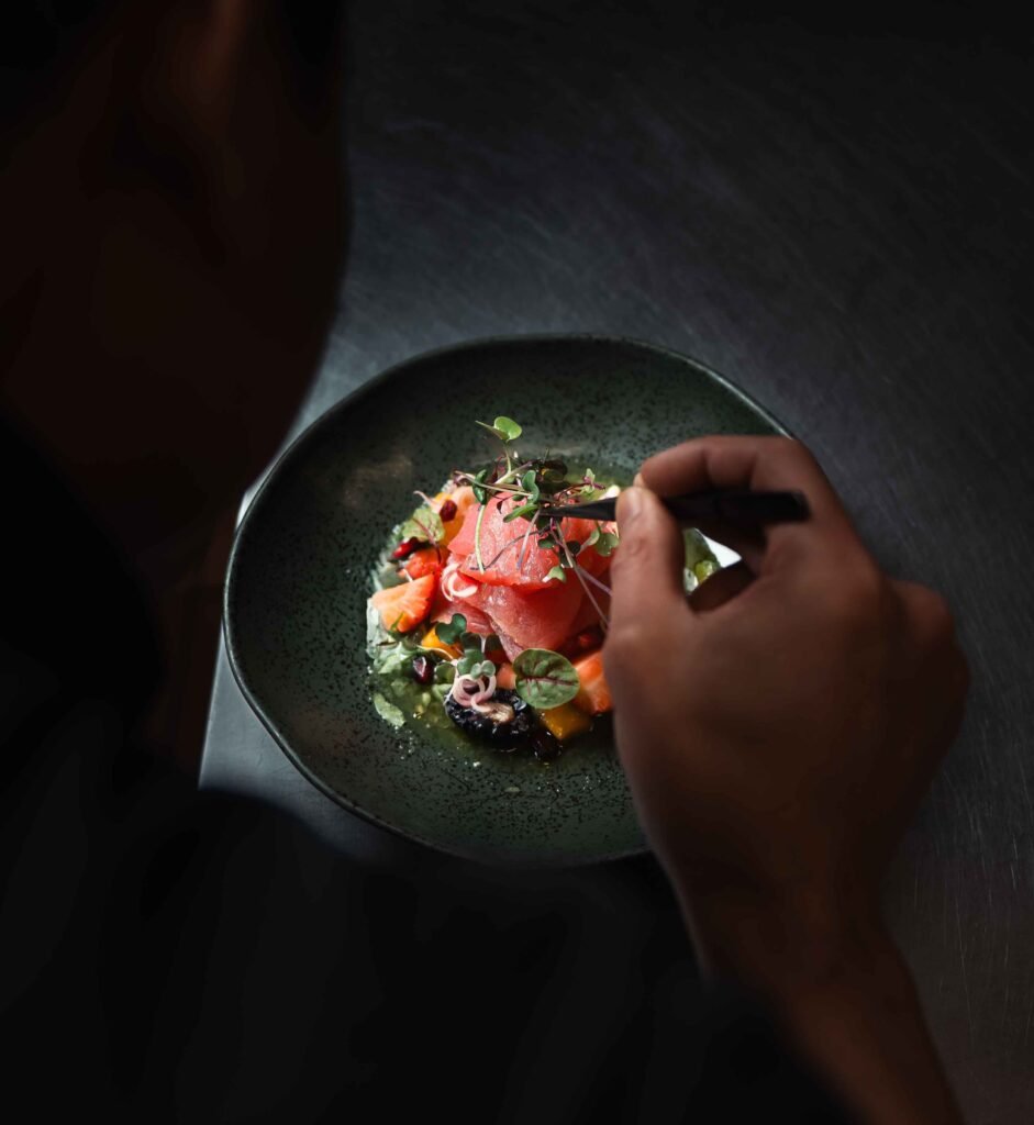 A food-photographer captures a person garnishing a gourmet dish with microgreens on a dark plate, featuring vibrant sliced fish and colorful vegetables artfully arranged against a dramatic background.