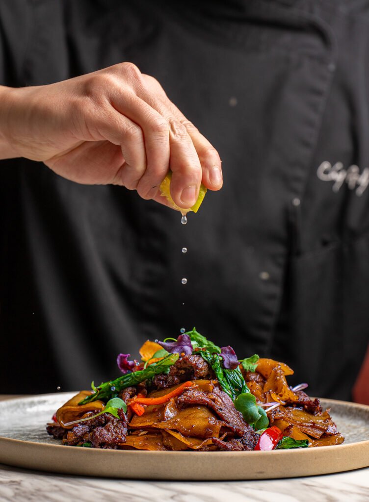 A person squeezes a slice of lemon over a plate of colorful sautéed vegetables and meat, garnished with greens, while wearing a black chef’s coat—captured by a skilled toronto-food-photographer.